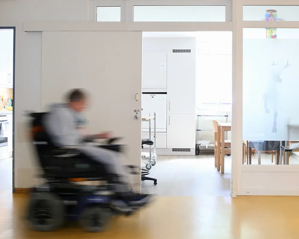 Ein Mann im Rollstuhl fährt den Flur entlang undan einer offenen Zimmertür vorbei