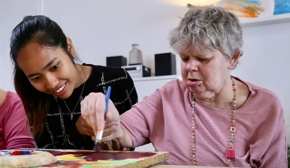 eine Frau mit Beeinträchtigung malt ein Bild und bekommt Unterstützung von einer Betreuerin