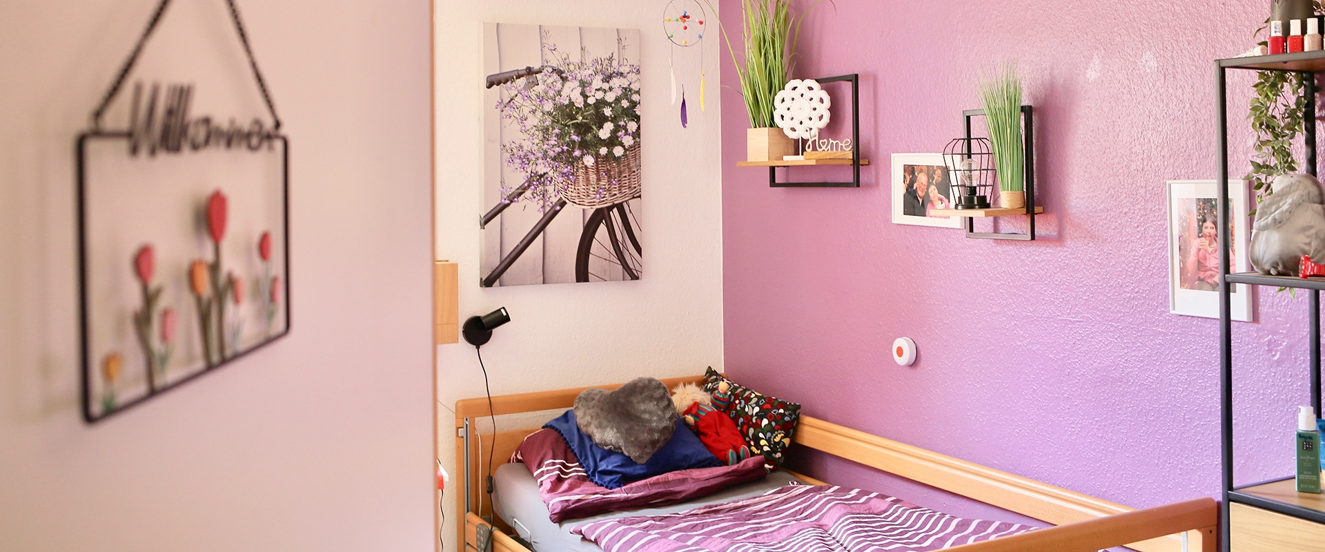 Blick in ein gemütliches Zimmer im betreuten Wohnen mit einer rosa Wand und einem bett mit Kuscheltieren.