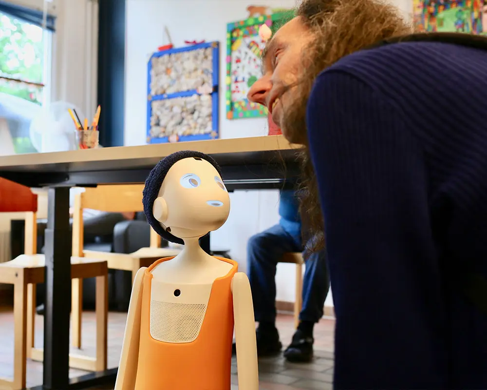 In einem Innenraum mit bunten Wandbildern steht eine kleine humanoide Roboterfigur auf dem Boden. Eine Person beugt sich nah zu dem Roboter hinunter.